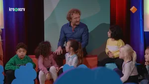 Een foto van een man met krullend haar. Hij zit voor een groot doek. Voor hem zitten zes kleine kinderen. Sommige van die kinderen kijken naar de man.