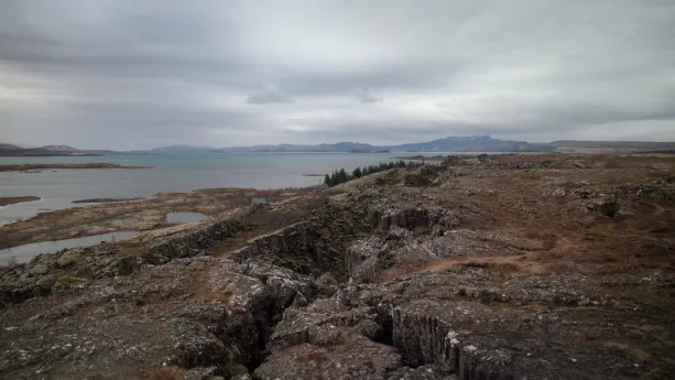 Een foto van een bergachtig landschap met veel kloven. Op de achtergrond is er een stuk bos te zien met erachter een grote plas water. In de verte zijn bergen te zien.