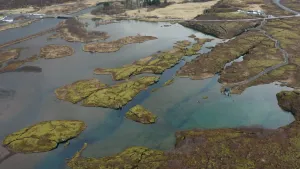 Een foto van een gebied waar kleine stukken groene grond liggen in helderblauw water. Door het water heen zijn lager gelegen stukken grond of bodem te zien. Achteraan is een weg te zien en aan twee kanten van de weg een klein groepje witte gebouwen.
