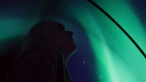 Een foto van onderzoeker Govert die naar boven kijkt naar de koepel boven zijn hoofd. Op de koepel is een animatie of video van een gedeelte van groen oplichtend poollicht in een donkere lucht met sterren te zien. 