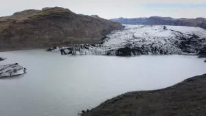 Een foto van een gebied met vooraan een stuk donkere grond, waar heel klein twee mensen te zien zijn. Achteraan is links een stenen gebergte te zien en rechtsachter een smeltende gletsjer van ijs met een zwarte kleur met een witte laag erop. 