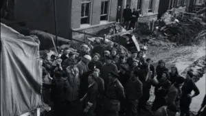 Een foto van de watersnoodramp van 1953. Je ziet een groep mensen bij een vrachtwagen staan. Op de achtergrond zie je een paar huizen. De straat is helemaal bedekt met modder. 
