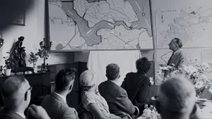 Een foto van een groep mensen in een kamer. Ze kijken allemaal naar grote landkaarten, van de provincie Zeeland, die aan de muur hangen. Een man, die voor de groep staat, wijst met een lange stok naar een punt op een van die landkaarten. 