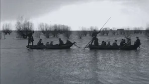 Een foto van de watersnoodramp van 1953. Je ziet een groot gebied met alleen maar water met daarin wat bomen. Je ziet ook twee roeiboten met daarin mensen. 