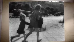 Een foto van een zwartwit-foto uit de Tweede Wereldoorlog. Je ziet daarop twee meisjes wegrennen. Ze hebben elkaars hand vast. Op de achtergrond zie je een straat met auto's.