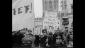 Een foto van een demonstratie in een stad. Je ziet mensen met spandoeken en borden met daarop teksten. De mensen dragen allemaal mondkapjes. Op een bord staat "Onze longen hun vuilnisvat". 