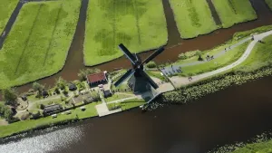 Een foto van een Nederlands landschap, gezien vanuit de lucht. Je ziet een molen, een huisje, weilanden, sloten en een paar wegen. Ook zie je mensen wandelen. 