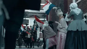 Een foto van een straat in een stad. Het speelt in de achttiende eeuw. In de verte komt er een koets aan. Erop staan mannen met pruiken en hoeden op. Langs de weg staan vrouwen met Nederlandse vlaggen te zwaaien. 