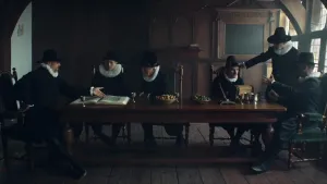 Een foto van zes mannen in zeventiende-eeuwse kleren. Ze zitten in een oud huis aan een lange houten tafel en zijn met elkaar in gesprek in twee groepen van drie man. De foto lijkt wel een zeventiende-eeuws schilderij.