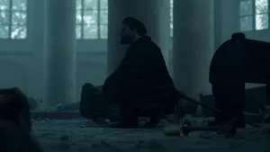 Een foto van een man die op zijn hurken zit, in een kerk. Het beeld is donker, maar je ziet een kerkbank, en allemaal stukken steen op de grond. Tussen de brokken steen ligt ook een kaarsenstandaard met de kaars ernaast. 