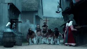 Een foto van een Middeleeuws uitziende straat. Aan de randen van de straat staan vrouwen. Midden op straat zie je vijf knielende mannen, met daarachter iemand van de kerk die een groot kruis in de lucht houdt. De mannen lijken te worden gestraft.