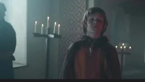 Een foto van een jonge jongen in een kamer van een kasteel. Hij heeft Middeleeuwse kleren aan. De jongen heeft zijn ogen dicht. Op de achtergrond zie je twee standaarden met brandende kaarsen erop. 