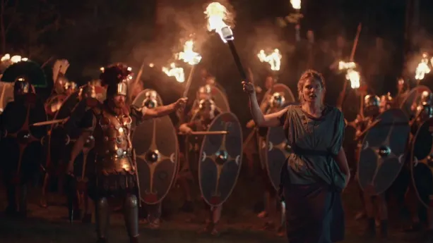 Een foto van een groep Romeinse soldaten die in het donker met wapens, schilden en fakkels staan. Voor hen staat er een generaal en een vrouw met beide een fakkel in hun handen.