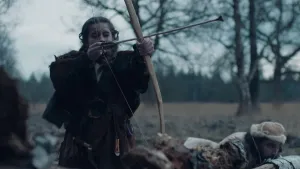 Een foto van een jonge vrouw in de prehistorie. Ze heeft een pijl en boog vast en richt die ergens op. Naast haar ligt een man met een bontmuts op de grond en hij loert ergens naar. 