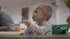 Een foto van een klein jongetje aan een tafel. Hij is aan het eten. Hij houdt zijn lepel omhoog en zegt iets. Naast hem zit nog iemand.