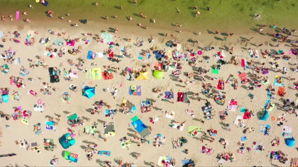 Een foto, vanuit de lucht genomen, van een strand vol met mensen. Boven in beeld zie je water dat groenig van kleur is. 