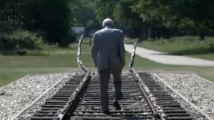 Een foto van nabestaande Salo Muller. Hij loopt tussen de spoorrails van het monument van Kamp Westerbork. Aan het eind gaan de spoorrails omhoog en zien ze er beschadigd uit.