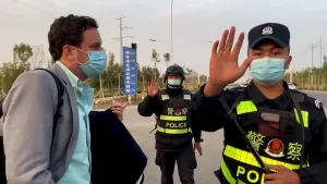 Een foto van de presentator. Hij heeft een mondkapje op en kijkt naar een Chinese politieagent. Die maakt een gebaar naar de camera dat er niet gefilmd mag worden. Op de achtergrond zie je een tweede politieagent die hetzelfde gebaar maakt.