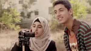 Een foto van een Palestijnse vrouw en man die naast elkaar staan. De vrouw houdt een video camera vast en kijkt op het beeldscherm van de camera.