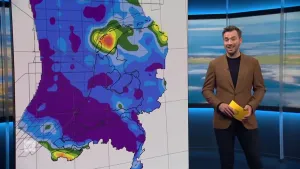 Een foto van een presentator die naast een kaart staat van Nederland en de Noordzee waarop de warmte van de aarde op te zien is. Vooral in een gebied in de Waddenzee is de aarde erg warm.