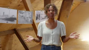 Een foto van een presentator die op een zolder staat met de armen gebogen met de handpalmen omhoog. Op de achtergrond hangen aan een lijn drie zwart-wit foto's: één van een grote boot in het water en twee van mensen bij woningen in een Molukse woonwijk.