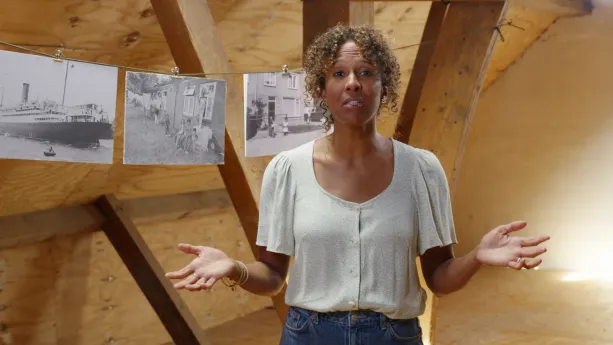Een foto van een presentator die op een zolder staat met de armen gebogen met de handpalmen omhoog. Op de achtergrond hangen aan een lijn drie zwart-wit foto's: één van een grote boot in het water en twee van mensen bij woningen in een Molukse woonwijk.