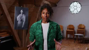 Een foto van een presentator die op een zolder staat. Op de achtergrond hangt een zwart-wit foto van Adolf Hitler leider van de nazi-partij in de Tweede Wereldoorlog aan een lijn. De presentator houdt de handen met gespreide vingers voor zich.