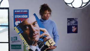 Een foto van een presentator voor een witte muur met ramen en politieke posters erop. De presentator heeft een poster vast van de politieke partij Lijst Pim Fortuyn. De foto en handtekening van Pim Fortuyn staan erop en er staat "at your service!" op. 
