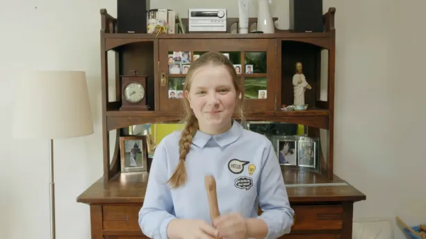 Een foto van Willemijn voor een bruine kast met foto's van Willemijn en haar familie en een beeldje van Jezus Christus erop, omdat Willemijn rooms-katholiek van geloof is. Willemijn heeft een houten blokfluit in haar handen en een lichtblauwe blouse aan.