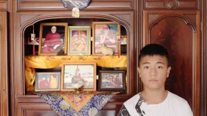 Een foto van Dawoe voor een bruine kast met foto's. Op de bovenste plank staan drie foto's van de boeddhistische spirituele leider de Dalai Lama op een gele doek, op een blauwe doek eronder drie foto's van boeddhistische monniken in rode of gele kleding.