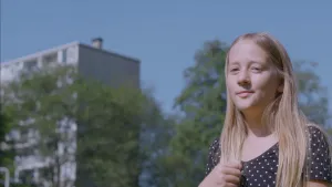 Een foto van Lune die buiten staat en rechtsvoor op de foto te zien is. Ze houdt haar hand in een vuist voor haar borstkas. Lune draagt een donkerkleurige blouse met witte stipjes erop. Linksachter is onscherp een flatgebouw te zien, ervoor groene bomen. 