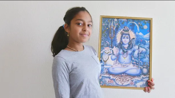 Een foto van Nandeni. Ze heeft een goudkleurige lijst vast met een kleurrijke tekening van de hindoeïstische god Shiva met een lichtblauwe huid, in kleermakerszit met een dier, een slang om de nek vóór een witte koe, bloemen, bergen en stromend water. 