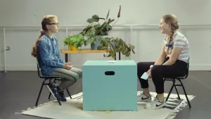 Een foto van Rouwayda, moslim, en Willemijn, katholiek, die op een groene stoel met zwarte poten zitten. Tussen hen in staat een lichtgroene, vierkante kist als tafel met een stapel papieren kaartjes erop. Willemijn heeft een kaartje vast. 