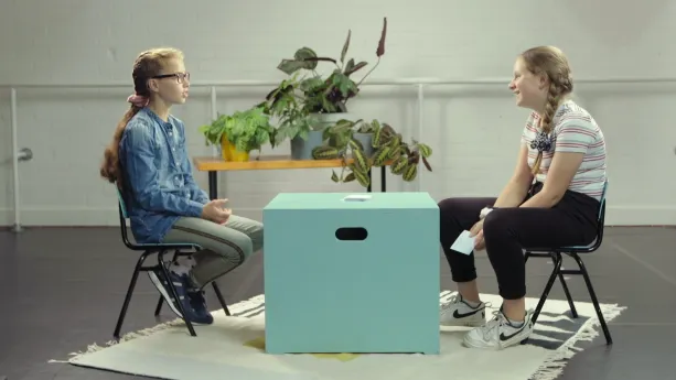 Een foto van Rouwayda, moslim, en Willemijn, katholiek, die op een groene stoel met zwarte poten zitten. Tussen hen in staat een lichtgroene, vierkante kist als tafel met een stapel papieren kaartjes erop. Willemijn heeft een kaartje vast. 