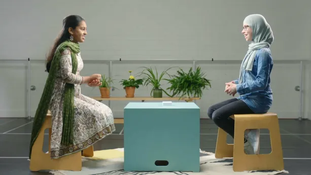 Een foto van Nandeni, hindoe, en Rouwayda, moslim met een hoofddoek, die op een houten kruk zitten op een wit vloerkleed met donkere lijnen en vormen. Tussen hen in staat een lichtgroene, vierkante kist als tafel met een stapel papieren kaartjes erop. 