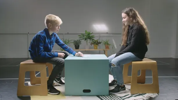 Een foto van Roeland, protestants, en Ronit, joods, die op een houten kruk zitten op een wit vloerkleed met grijze lijnen en vormen. Tussen hen in staat een lichtgroene, vierkante kist als tafel met een stapel papieren kaartjes erop. Roeland pakt er één.