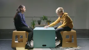 Een foto van Nandeni, hindoe en Roelof, protestants, die op een houten kruk zitten op een wit vloerkleed met donkere lijnen en vormen. Tussen hen in staat een lichtgroene, vierkante kist als tafel met een stapel papieren kaartjes erop. Roelof pakt er één.