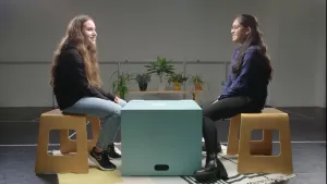 Een foto van Ronit, joods, en Nandeni, hindoe, die op een houten kruk zitten op een wit vloerkleed met donkere lijnen en vormen. Tussen hen in staat een lichtgroene, vierkante kist als tafel. Er liggen drie stapels papieren kaartjes op. 