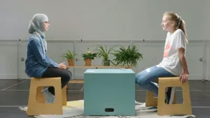 Een foto van Rouwayda, moslim met hoofddoek, en Lune, humanist, die op een houten kruk zitten op een wit vloerkleed met donkere lijnen en vormen. Tussen hen in staat een lichtgroene, vierkante kist als tafel. Er liggen twee stapels papieren kaartjes op. 