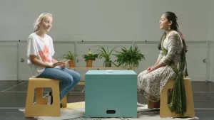 Een foto van Lune, humanist, en Nandeni, hindoe, die op een houten kruk zitten op een wit vloerkleed met donkere lijnen en vormen. Tussen hen in staat een lichtgroene, vierkante kist als tafel. Er ligt een stapel papieren kaartjes op. 