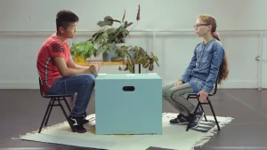 Een foto van Dawoe, boeddhist, en Rouwayda, moslim, die op een groene stoel met zwarte poten zitten op een wit vloerkleed met donkere vormen erop. Tussen hen in staat een lichtgroene, vierkante kist als tafel. Er liggen drie stapels papieren kaartjes op. 