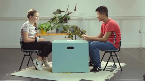 Een foto van Willemijn, katholiek, en Dawoe, boeddhist, die op een groene stoel met zwarte poten zitten op een wit vloerkleed. Tussen hen in staat een lichtgroene, vierkante kist als tafel. Er ligt een stapel papieren kaartjes op. Dawoe heeft er één vast.