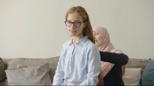 Een foto van Rouwayda in een lichtblauw en wit gestreepte blouse. Haar moeder zit erachter op een grijze bank en raakt het haar van Rouwayda aan de achterkant aan. De moeder van Rouwayda heeft een bruinroze hoofddoek om, omdat ze moslim is van geloof.