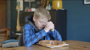 Een foto van Roeland die aan tafel zit. Hij heeft zijn handen gevouwen met de ellebogen op tafel en heeft zijn ogen dicht. Hij is protestants van geloof en is aan het bidden. Voor hem op tafel staat een wit bord met een pizza met stukjes groenten erop. 