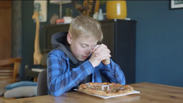 Een foto van Roeland die aan tafel zit. Hij heeft zijn handen gevouwen met de ellebogen op tafel en heeft zijn ogen dicht. Hij is protestants van geloof en is aan het bidden. Voor hem op tafel staat een wit bord met een pizza met stukjes groenten erop.