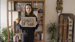 Een foto van Ronit voor een kast met boeken en foto's. Ronit is joods en heeft in haar handen een zwart-wit foto met een familie erop, waarschijnlijk haar joodse voorouders. Rechts staan drie stroken van stof met kleurige vormen erop in een lijst.