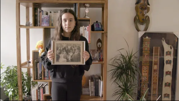 Een foto van Ronit voor een kast met boeken en foto's. Ronit is joods en heeft in haar handen een zwart-wit foto met een familie erop, waarschijnlijk haar joodse voorouders. Rechts staan drie stroken van stof met kleurige vormen erop in een lijst.