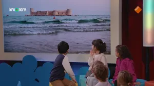 Een foto van zes kleine kinderen die naar een groot scherm kijken. Op dat scherm zie je een zee met golven. In de zee zie je een eiland met daarop een groot kasteel. 