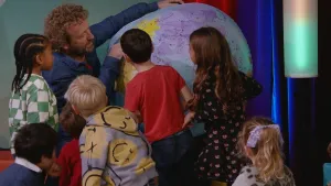 Een foto van een man met zes kleine kinderen. De man houdt een opblaasbal in de vorm van de Aarde vast. De man wijst naar een land op die Aarde. De kinderen kijken aandachtig naar dat land.