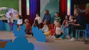 Een foto van een studio met daarin twee mannen en een groep kleine kinderen. Ze kijken naar een groot scherm. Daarop zie je bloemen. Een van de mannen speelt gitaar.